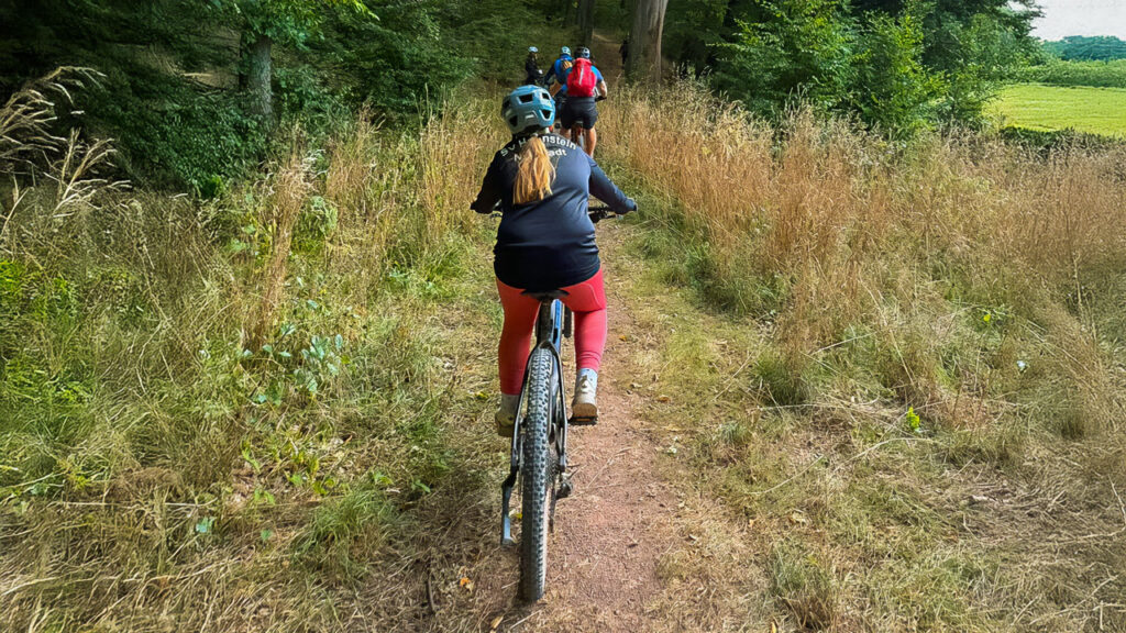Radgruppe auf schmalem Waldpfad; im Vordergrund eine Radlerin mit Helm und roten Leggings, vor ihr mehrere Mitfahrende – Sommergrün am Wegesrand.