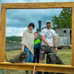 Familie mit Kind und zwei Hunden posiert im hölzernen Selfie-Rahmen „Neustadt/Harz“ am Dampfloksteig; im Hintergrund Holzeisenbahn-Wagen, Hügel und wolkiger Himmel, QR-Codes am Rahmen sichtbar.