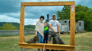 Familie mit Kind und zwei Hunden posiert im hölzernen Selfie-Rahmen „Neustadt/Harz“ am Dampfloksteig; im Hintergrund Holzeisenbahn-Wagen, Hügel und wolkiger Himmel, QR-Codes am Rahmen sichtbar.