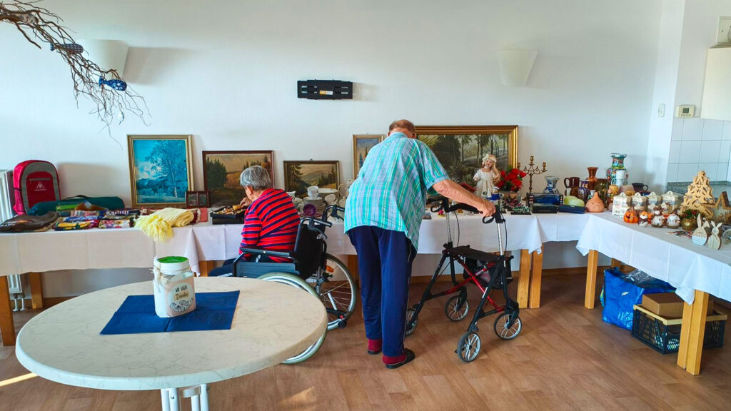 Zwei Senioren stöbern an einer langen Flohmarkt-Tafel im Aufenthaltsraum; eine Person im Rollstuhl, eine mit Rollator. Auf den Tischen Gemälde, Keramik und Dekostücke; Spendenglas im Vordergrund.
