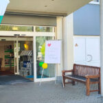 Gebäudeeingang mit offenen Schiebetüren: zwei grün-weiße Beachflags ‚Kompetenz in Pflege‘, Willkommensschild ‚Garagen-Flohmarkt‘ mit Ballons, Sitzbänke neben dem Eingang.