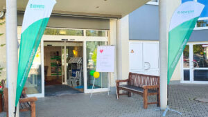 Gebäudeeingang mit offenen Schiebetüren: zwei grün-weiße Beachflags ‚Kompetenz in Pflege‘, Willkommensschild ‚Garagen-Flohmarkt‘ mit Ballons, Sitzbänke neben dem Eingang.