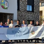 Gruppenfoto vor einem modernen Seniorenwerk-Gebäude: zwölf Mitarbeitende und Auszubildende halten ein großes Banner mit der Aufschrift „Wir leben Pflege.“; rechts steht ein weißes Dienstfahrzeug, sonniger Tag.