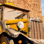 Historischer gelber Oldtimer-Bus mit schwarzem Dach und großen Rundscheinwerfern, aufgenommen vor einer alten Natursteinmauer bei Sonnenschein.