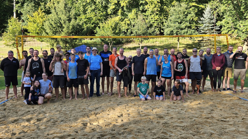 Gruppenfoto der Teilnehmenden des Beachvolleyball-Turniers auf dem Sandfeld, lächelnd vor gelbem Netz; Erwachsene und Kinder, grüner Park im Hintergrund.