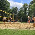Beachvolleyball auf dem Sandfeld: gemischtes Team im Spiel, Spieler rechts baggert den Ball; Sonnenschein, Bäume und Zuschauerzelte im Hintergrund.
