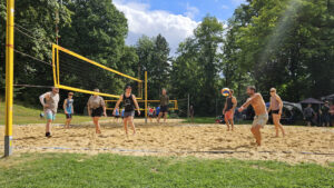 Beachvolleyball auf dem Sandfeld: gemischtes Team im Spiel, Spieler rechts baggert den Ball; Sonnenschein, Bäume und Zuschauerzelte im Hintergrund.