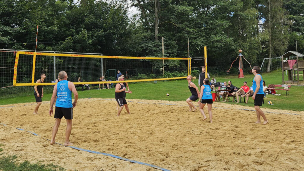 Ballwechsel beim Beachvolleyball: Teams in schwarzen und blauen Trikots, Annahme am Netz; Zuschauer auf Bänken, Spielplatzskulptur und Bäume dahinter.
