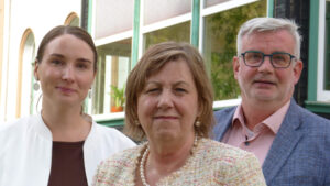 Pressefoto beim 19. Thüringer Pflegesymposium in Ilfeld-Harztor: drei Personen im Halbportrait vor grünen Fensterrahmen – mittig Frau mit Perlenkette, links Frau in weißer Jacke, rechts Mann mit Brille.