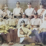 Historisches, handkoloriertes Gruppenfoto: rund ein Dutzend uniformierte Feuerwehrmänner in hellen Jacken und Helmen, mit Lederkoppeln, Äxten und Werkzeugen; Studioaufnahme – einige sitzen, andere stehen, einer liegt vorn auf einem Teppich (um 1900).