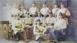 Historisches, handkoloriertes Gruppenfoto: rund ein Dutzend uniformierte Feuerwehrmänner in hellen Jacken und Helmen, mit Lederkoppeln, Äxten und Werkzeugen; Studioaufnahme – einige sitzen, andere stehen, einer liegt vorn auf einem Teppich (um 1900).