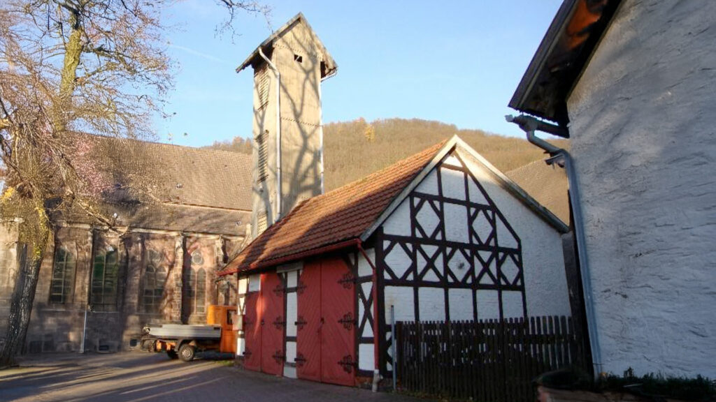 	1.	Altes Feuerwehrhaus in Fachwerkbauweise mit roten Doppeltoren und hohem Schlauchtrockenturm; links die Kirche, Herbstsonne wirft Baumschatten, kleiner Kommunalkipper im Hof.