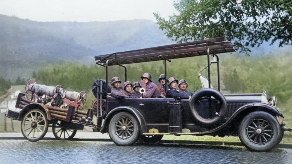 Historische Aufnahme: offener Feuerwehr-Wagen der 1930er Jahre mit acht Feuerwehrmännern in Helmen; seitlich Ersatzrad und Leitergestell. Hinten angehängt ein Schlauchwagen mit aufgewickelten Schläuchen. Ländliche Kulisse mit Hügeln, am Ufer eines Teichs.