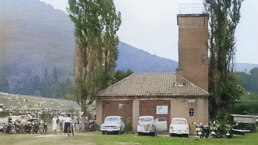 Historische Aufnahme eines kleinen Feuerwehrgerätehauses mit Schlauchtrockenturm; davor parken 1950er/60er-Jahre Pkw und mehrere Motorräder, links eine Gruppe Menschen, im Hintergrund Wiese und bewaldete Hügel.