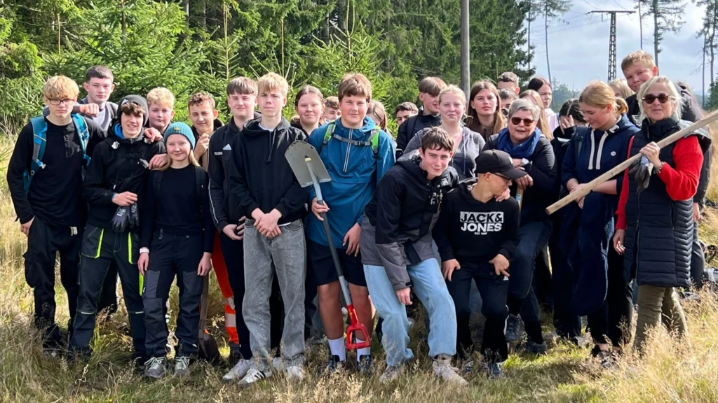 Große Gruppe Jugendlicher mit Begleitpersonen auf einer Lichtung; ein Schüler hält einen Spaten, im Hintergrund dichter Nadelwald.