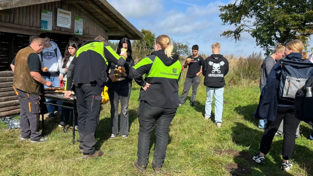 Pause an einer Schutzhütte: Forstleute grillen, Jugendliche stehen in kleinen Gruppen zusammen; sonniger Moment auf der Wiese.