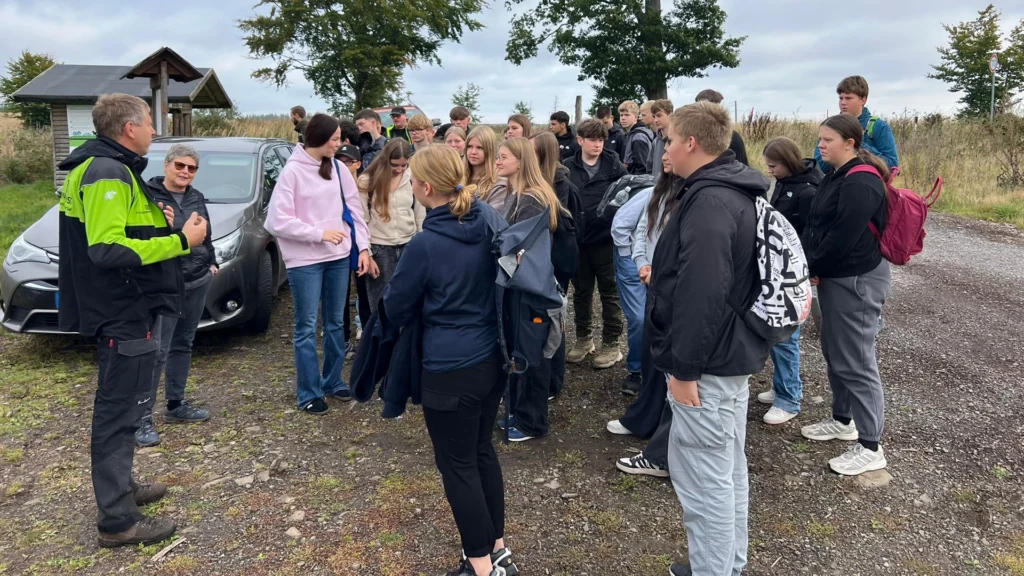 Forstmitarbeiter gibt einer großen Schülergruppe eine Einweisung auf einem Waldparkplatz; Rucksäcke und Wetterkleidung, bedeckter Himmel.
