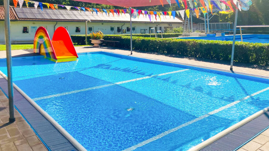 Neues Kinder-Planschbecken im Waldbad Ilfeld: klares Wasser, Fontänen und Sonnensegel; links die bunte Regenbogen-Rutsche, Wimpelketten darüber.