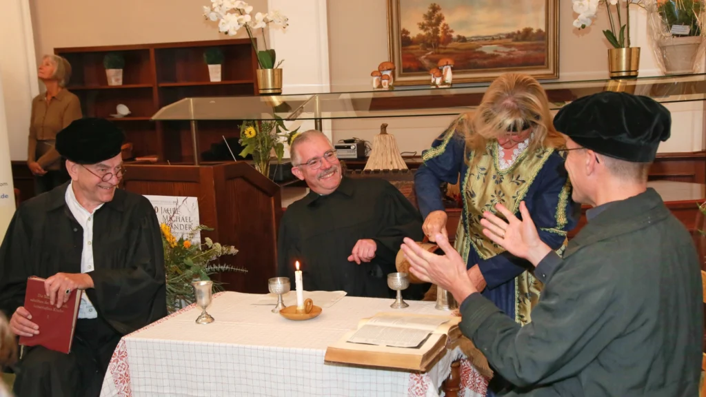 Szenische Darstellung am Tisch: vier Personen in historischen Kostümen mit Kerze, Kelchen und aufgeschlagenem Buch; heitere Runde im Saal.