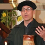 Darsteller in schwarzer Gelehrtenrobe und Barett gestikuliert beim Vortrag; Kerze und Tonkrug im Vordergrund, Blumenarrangement dahinter.