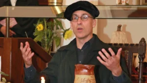Darsteller in schwarzer Gelehrtenrobe und Barett gestikuliert beim Vortrag; Kerze und Tonkrug im Vordergrund, Blumenarrangement dahinter.