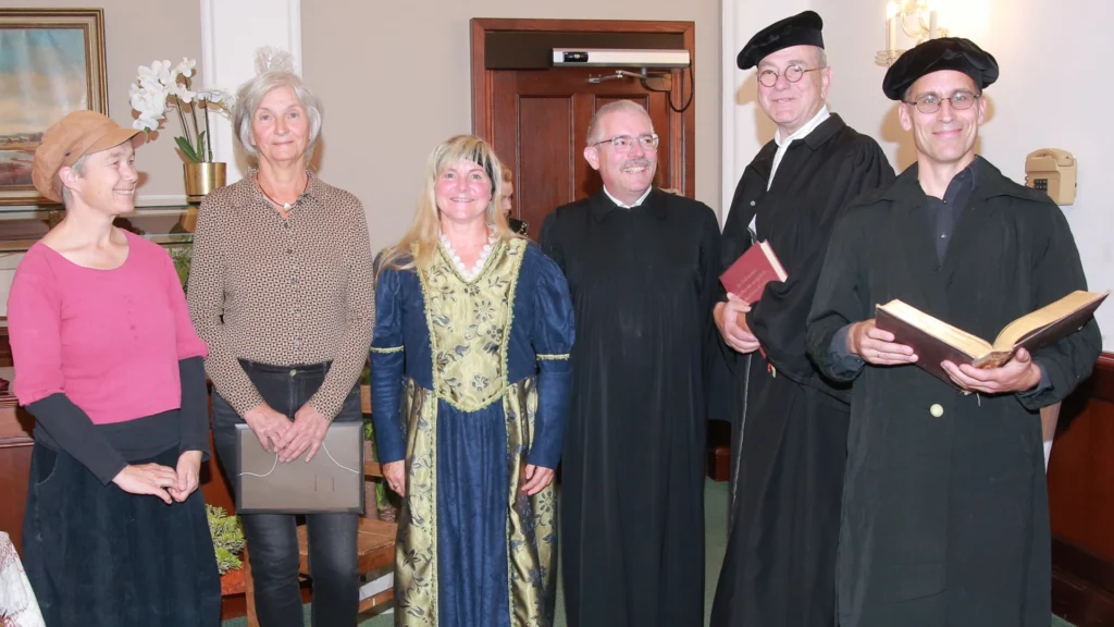 Gruppe von Mitwirkenden: zwei Frauen in Alltagskleidung, eine Frau im Renaissance-Kleid, drei Männer in schwarzen Gelehrtenroben mit Büchern, Innenraum.
