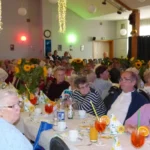 Seniorenfest Harztor: voll besetzter Saal mit langen, festlich gedeckten Tischen, Sonnenblumen-Deko und Getränken; Gäste verfolgen das Programm.