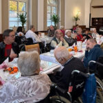 Bewohnerinnen und Bewohner eines Pflegeheims sitzen in einem festlich gedeckten Speisesaal an langen Tischen und genießen ein gemeinsames Herbstfest. Einige Gäste sitzen in Rollstühlen, andere unterhalten sich oder klatschen Beifall. Auf den Tischen stehen kleine Kürbisse und herbstliche Dekorationen, im Hintergrund spielt ein Musiker. Die Atmosphäre wirkt warm und gesellig.