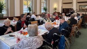 Bewohnerinnen und Bewohner eines Pflegeheims sitzen in einem festlich gedeckten Speisesaal an langen Tischen und genießen ein gemeinsames Herbstfest. Einige Gäste sitzen in Rollstühlen, andere unterhalten sich oder klatschen Beifall. Auf den Tischen stehen kleine Kürbisse und herbstliche Dekorationen, im Hintergrund spielt ein Musiker. Die Atmosphäre wirkt warm und gesellig.