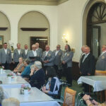 Ein Männerchor in grauen Sakkos singt in der Aula der Neanderklinik Harzwald vor zahlreichen Seniorinnen und Senioren. Die Zuhörer sitzen an festlich gedeckten Tischen, während die Sänger in Reihen stehen und aus Notenmappen vortragen. Die Stimmung wirkt konzentriert und herzlich.