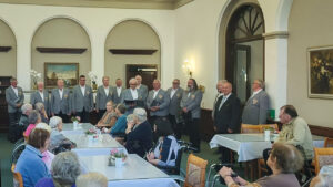 Ein Männerchor in grauen Sakkos singt in der Aula der Neanderklinik Harzwald vor zahlreichen Seniorinnen und Senioren. Die Zuhörer sitzen an festlich gedeckten Tischen, während die Sänger in Reihen stehen und aus Notenmappen vortragen. Die Stimmung wirkt konzentriert und herzlich.