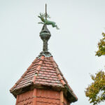 Spitzes Türmchen mit roten Dachziegeln und einer Hirschfigur als Wetterfahne auf der Spitze, daneben ragen grüne Äste eines Baumes in den hellgrauen Himmel.