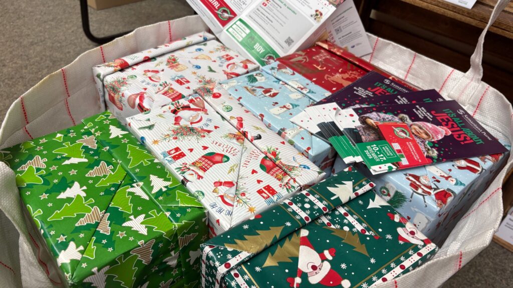 Ein großer Stoffbeutel voller bunt verpackter Schuhkartons, die für die Aktion „Weihnachten im Schuhkarton“ vorbereitet wurden. Auf den Kartons sind weihnachtliche Motive wie Weihnachtsmänner, Schneemänner und Tannenbäume zu sehen. Obenauf liegen Flyer der Aktion mit Hinweisen zum Packen der Geschenke.