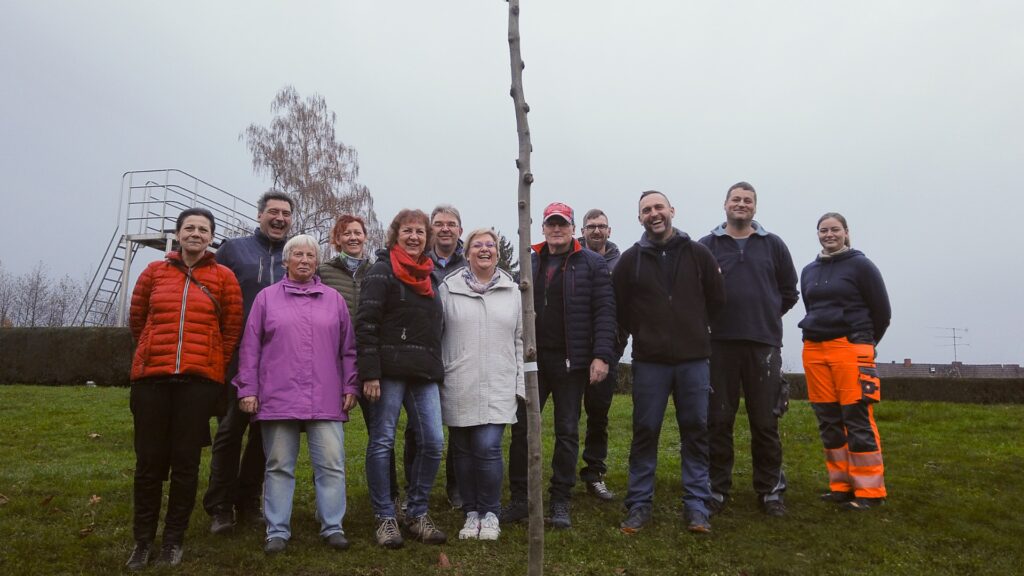 Zehn Mitglieder des Arreé-Traditionsvereins und des Ortschaftsrates pflanzen gemeinsam einen jungen Lederhülsenbaum auf der grünen Liegewiese des Freibads Niedersachswerfen. Im Hintergrund sind die leeren Edelstahlbecken des Bades zu sehen.