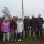 Zehn Mitglieder des Arreé-Traditionsvereins und des Ortschaftsrates pflanzen gemeinsam einen jungen Lederhülsenbaum auf der grünen Liegewiese des Freibads Niedersachswerfen. Im Hintergrund sind die leeren Edelstahlbecken des Bades zu sehen.