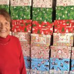 Frau Christine Andrae aus Ilfeld steht lächelnd vor einer Wand aus zahlreichen bunt verpackten Schuhkartons. Sie organisiert seit vielen Jahren die Aktion „Weihnachten im Schuhkarton“ in der Gemeinde Harztor und hat auch in diesem Jahr wieder rund 200 Geschenkboxen liebevoll vorbereitet.