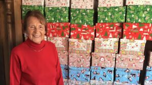 Frau Christine Andrae aus Ilfeld steht lächelnd vor einer Wand aus zahlreichen bunt verpackten Schuhkartons. Sie organisiert seit vielen Jahren die Aktion „Weihnachten im Schuhkarton“ in der Gemeinde Harztor und hat auch in diesem Jahr wieder rund 200 Geschenkboxen liebevoll vorbereitet.