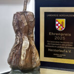 Eine bronzefarbene, abstrakte Skulptur steht neben einer schwarzen Gedenkplakette. Die Plakette zeigt das Wappen des Landkreises Nordhausen und die Inschrift "Ehrenpreis 2025" für die Organisation des größten Volkslaufes.