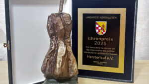 Eine bronzefarbene, abstrakte Skulptur steht neben einer schwarzen Gedenkplakette. Die Plakette zeigt das Wappen des Landkreises Nordhausen und die Inschrift "Ehrenpreis 2025" für die Organisation des größten Volkslaufes.