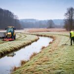 Pflegemaßnahmen im Brandesbachtal bei Ilfeld: Zwei Landschaftspfleger in Warnwesten mähen und beräumen eine feuchte Wiesenfläche entlang eines kleinen Baches. Im Hintergrund sind sanfte Hügel und winterlich kahle Bäume zu sehen, vorn steht ein kleiner Traktor mit Anhänger für das Mähgut. Die Szene zeigt eine schonende, naturnahe Landschaftspflege unter bedecktem Himmel. KI