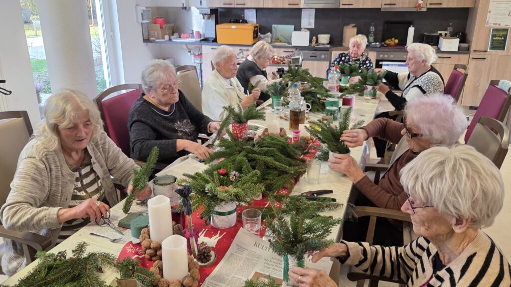 Eine Gruppe von sieben älteren Frauen sitzt an einem Tisch und bindet gemeinsam Adventskränze aus Tannenzweigen. Auf dem Tisch liegen Zeitungen, Gartenscheren, Tannengrün und dekorative Nüsse.