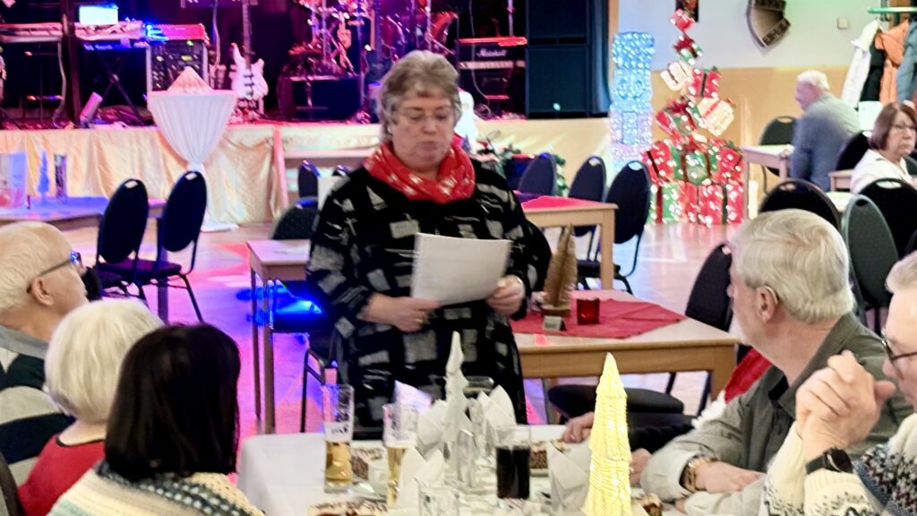 Die Vorsitzende des VdK Ortsverbandes Ilfeld hält in einem festlich dekorierten Saal, vor einer beleuchteten Bühne, eine Rede vor den anwesenden Mitgliedern. Die Rednerin trägt eine schwarze Bluse mit weißem Muster und einen roten Schal.