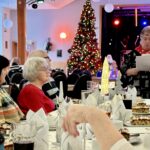 Nahaufnahme eines Tisches auf der VdK Weihnachtsfeier, an dem Mitglieder aufmerksam der Rede der Vorsitzenden zuhören. Der Tisch ist mit kleinen Weihnachtsbäumen, Gläsern und Kuchen gedeckt.