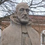 Nahaufnahme einer verwitterten Steinstatue eines Mannes mit Bart und Halskrause, die Michael Neander darstellt. Im Hintergrund sind kahle Bäume und die Fassade eines historischen Gebäudes aus rotem Backstein mit Bogenfenstern zu sehen.