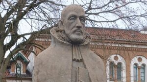 Nahaufnahme einer verwitterten Steinstatue eines Mannes mit Bart und Halskrause, die Michael Neander darstellt. Im Hintergrund sind kahle Bäume und die Fassade eines historischen Gebäudes aus rotem Backstein mit Bogenfenstern zu sehen.