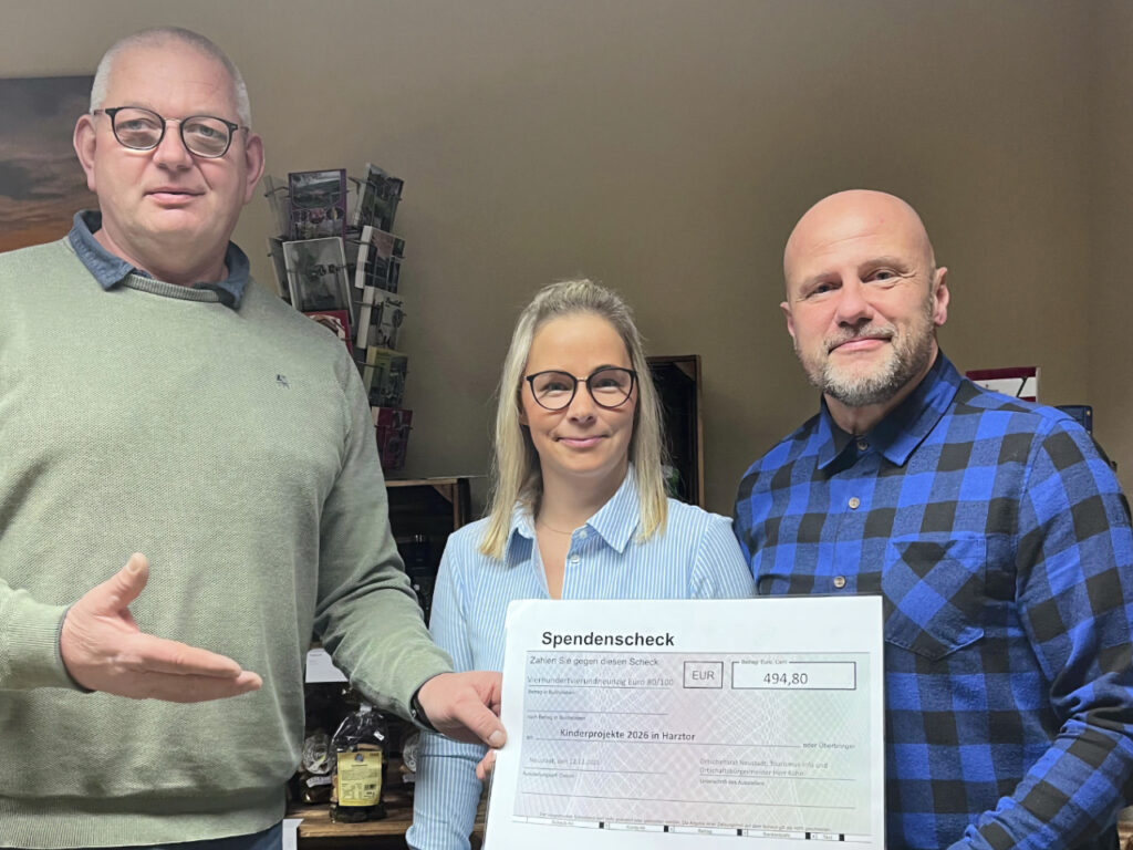 Drei Personen stehen nebeneinander und halten gemeinsam einen großen symbolischen Spendenscheck in die Kamera. Links steht ein Mann in einem grünen Pullover, in der Mitte eine Frau mit Brille und hellblau gestreifter Bluse, und rechts ein Mann im blau-karierten Hemd. Auf dem Scheck steht die Summe von 494,80 Euro mit dem Verwendungszweck „Kinderprojekte 2026 in Harztor“.