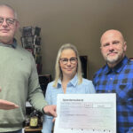 Drei Personen stehen nebeneinander und halten gemeinsam einen großen symbolischen Spendenscheck in die Kamera. Links steht ein Mann in einem grünen Pullover, in der Mitte eine Frau mit Brille und hellblau gestreifter Bluse, und rechts ein Mann im blau-karierten Hemd. Auf dem Scheck steht die Summe von 494,80 Euro mit dem Verwendungszweck „Kinderprojekte 2026 in Harztor“.