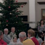 Blick in einen Saal mit einem großen, geschmückten Weihnachtsbaum und Senioren an Kaffeetafeln. Rechts im Bild sorgen ein Musiker am Keyboard und eine Sängerin für die musikalische Umrahmung.