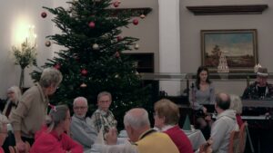 Blick in einen Saal mit einem großen, geschmückten Weihnachtsbaum und Senioren an Kaffeetafeln. Rechts im Bild sorgen ein Musiker am Keyboard und eine Sängerin für die musikalische Umrahmung.