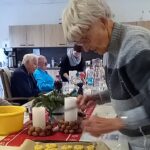 Mehrere Senioren sitzen in der Tagespflege an einem Tisch und bereiten Plätzchen zu. Eine Frau bedient einen Fleischwolf, eine andere verziert die ausgestochenen Plätzchen auf dem Blech. Auf dem Tisch stehen Kerzen und weihnachtliche Dekoration.